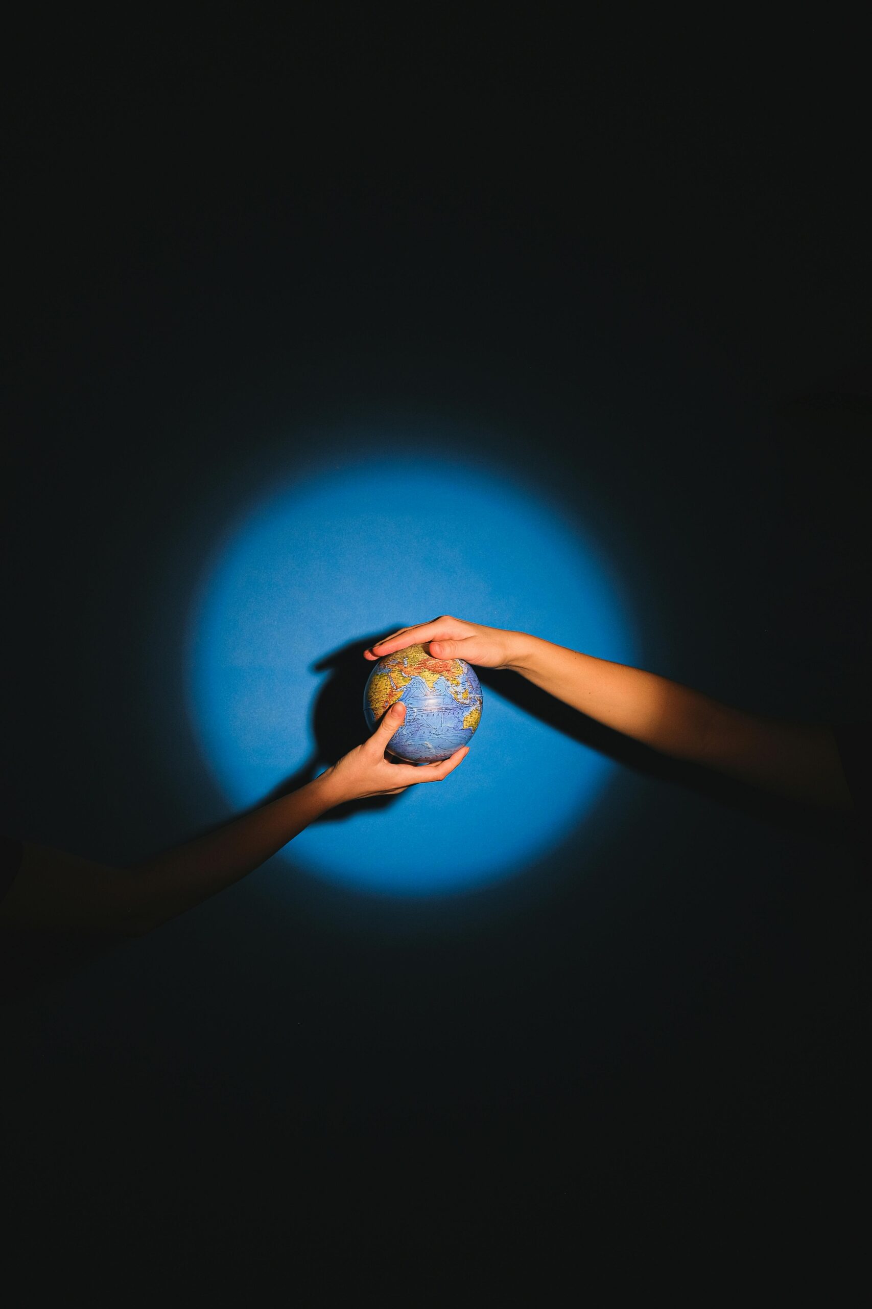 Two hands exchange a glowing globe with spotlight in a dark setting, symbolizing connection.